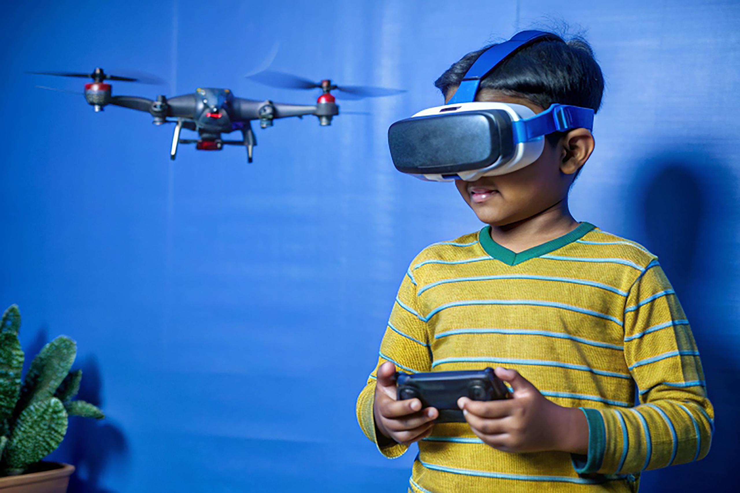 Intelligent indian kid controlling drone by wearing VR or virtual reality headset and controller at home - concept of modern technology